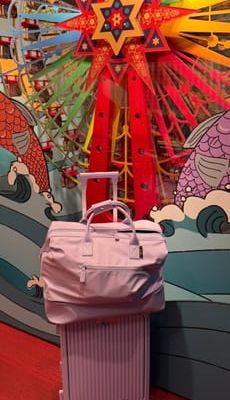 Customer-uploaded image. Click to see more details. The image represents: A pink rolling suitcase with a matching pink travel bag placed on top, positioned in front of a colorful decorative star-shaped backdrop with red, yellow, and green rays.