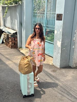 Customer-uploaded image. Click to see more details. The image represents: A woman wearing sunglasses and a floral dress stands with a light blue suitcase and gold bag outside a building with blue-green gates and walls.