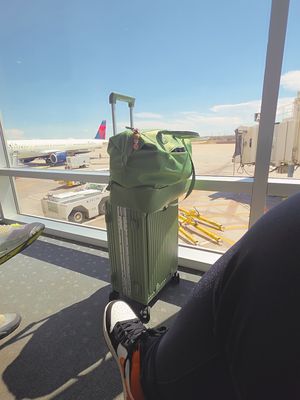 Customer-uploaded image. Click to see more details. The image represents: A green rolling suitcase with a garment bag draped over it sits in an airport terminal by a window, with an airplane visible on the tarmac outside and airport ground equipment in the background.