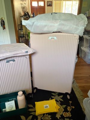 Customer-uploaded image. Click to see more details. The image represents: Two pink rolling suitcases in a home interior, one with a white garment bag draped over it, surrounded by packing materials on a patterned rug.