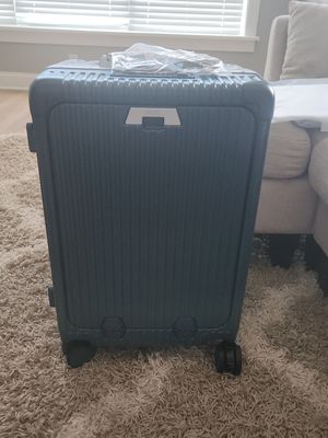 Customer-uploaded image. Click to see more details. The image represents: A gray hard-shell rolling suitcase with vertical ribbed texture, sitting on a beige carpet in a bright bedroom. The suitcase has black wheels and a handle, with plastic-wrapped items on top. A white chair is visible in the background near a window with blinds.