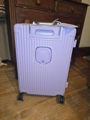 Customer-uploaded image. Click to see more details. The image represents: A lavender-colored hard-shell rolling suitcase with vertical ribbed texture, four black wheels, and a retractable handle, positioned in front of a dark wooden dresser on a wooden floor.