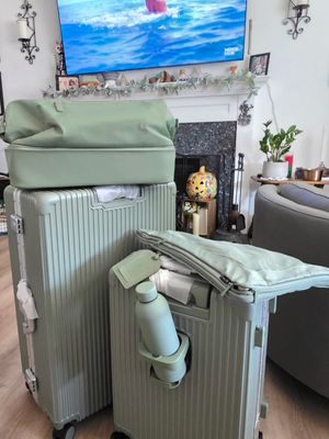 Customer-uploaded image. Click to see more details. The image represents: A living room with a mounted flat-screen TV displaying a beach scene, a green armchair, decorative wall sconces, a fireplace mantel with plants and photos, and two light green rolling suitcases in the foreground on a wooden floor.