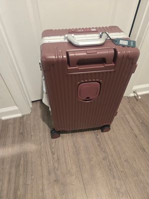 Customer-uploaded image. Click to see more details. The image represents: A burgundy hardshell rolling suitcase with a ribbed texture stands on a wooden floor against a white wall. It features a top handle, front pocket, and four wheels.